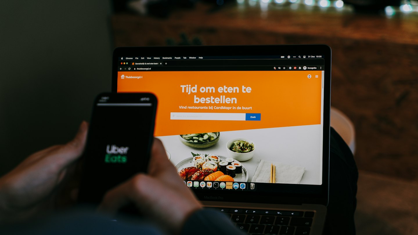 Dłonie trzymają smartfon z logo Uber Eats obok laptopa.